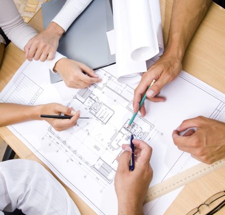 Baubesprechung Tisch mit Architektenplan und Händen die darauf zeigen oder schreiben. Stellt eine Baubesprechung dar.